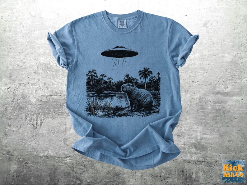 May include: A light blue t-shirt featuring a black and white graphic of a capybara standing near a body of water under a hovering UFO. The design includes palm trees and other foliage. The shirt has a crew neck and short sleeves.