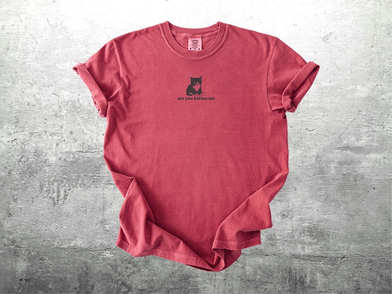May include: A red t-shirt with rolled-up sleeves. The shirt features a small black graphic of a cat and the phrase "are you kitten me" printed below the graphic. The shirt is displayed on a gray surface.
