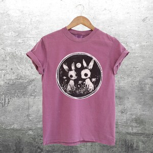 Op de afbeelding: Een roze T-shirt met een zwart-witte afbeelding van twee konijnen in een cirkel. De konijnen hebben grote ogen en zijn omringd door bloemen.