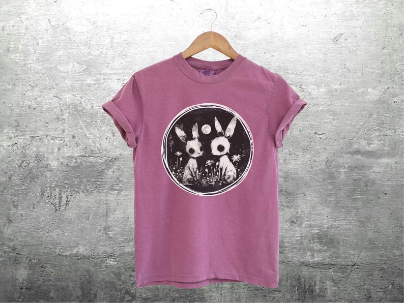 Op de afbeelding: Een roze T-shirt met een zwart-witte afbeelding van twee konijnen in een cirkel. De konijnen hebben grote ogen en zijn omringd door bloemen.