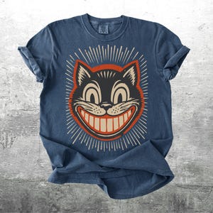 Op de afbeelding: Een donkerblauw t-shirt met ronde hals en een grafische print van een zwart kattengezicht met grote ogen en een brede grijns. De kat is omlijst door een rode cirkel en witte stralen. De tekst Kick Ash Co staat in de rechterbenedenhoek.