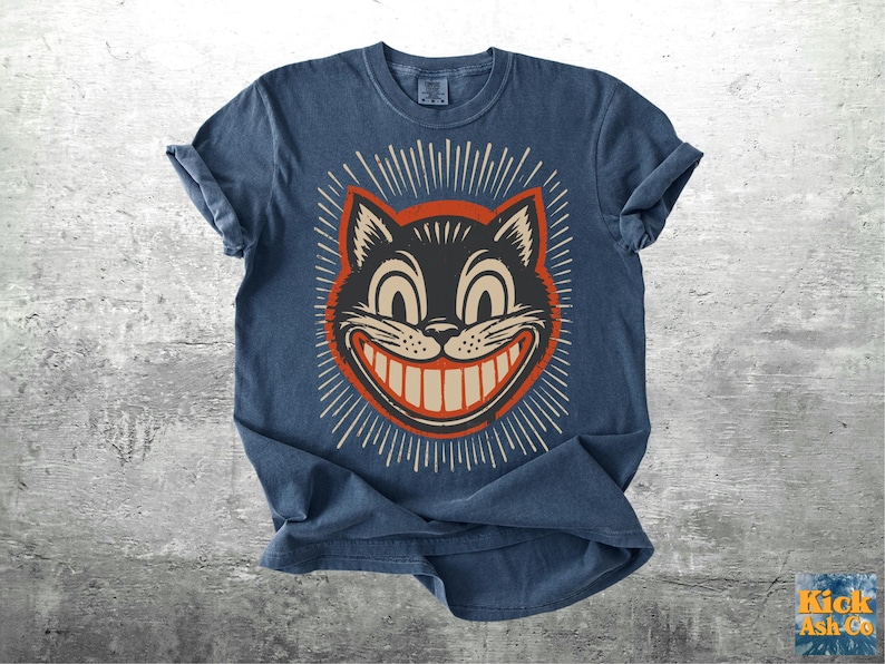 Op de afbeelding: Een donkerblauw t-shirt met ronde hals en een grafische print van een zwart kattengezicht met grote ogen en een brede grijns. De kat is omlijst door een rode cirkel en witte stralen. De tekst Kick Ash Co staat in de rechterbenedenhoek.