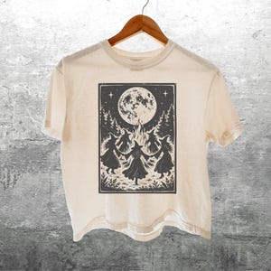 May include: A cream-colored short-sleeve t-shirt featuring a black graphic print of four witches dancing in a circle around a bonfire under a large full moon, surrounded by pine trees and stars, all contained within a rectangular border.