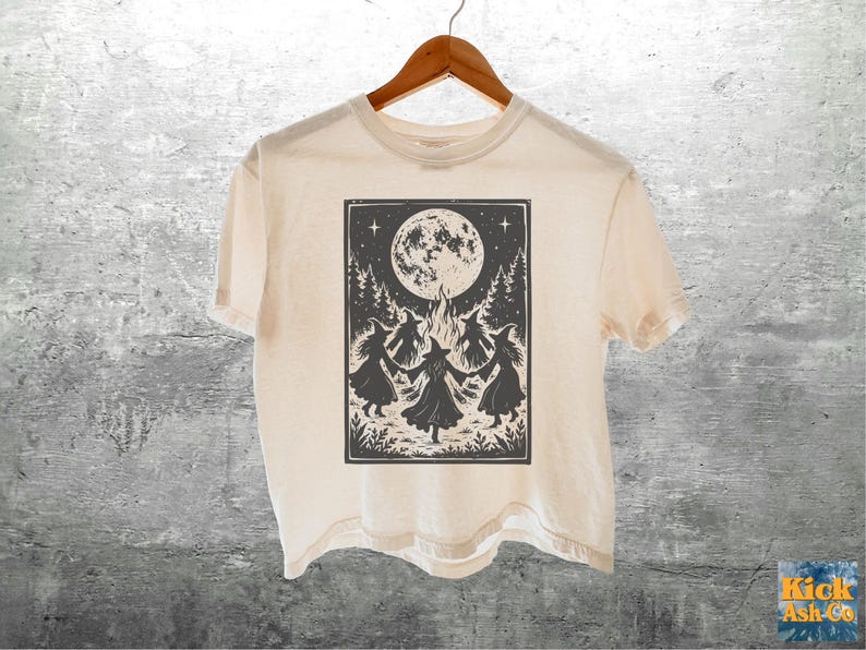 May include: A cream-colored short-sleeve t-shirt featuring a black graphic print of four witches dancing in a circle around a bonfire under a large full moon, surrounded by pine trees and stars, all contained within a rectangular border.