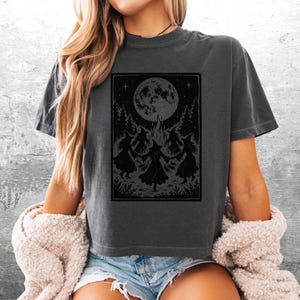 May include: A charcoal gray short-sleeve t-shirt featuring a black graphic print of three figures in pointed hats dancing in a circle around a campfire under a large full moon, surrounded by trees and foliage in a rectangular frame.