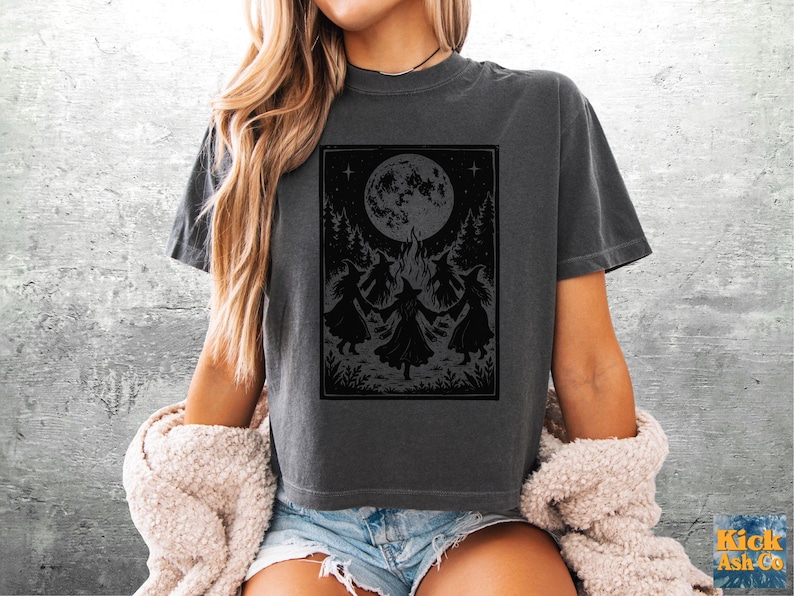 May include: A charcoal gray short-sleeve t-shirt featuring a black graphic print of three figures in pointed hats dancing in a circle around a campfire under a large full moon, surrounded by trees and foliage in a rectangular frame.
