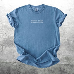 May include: A blue t-shirt with the phrase "rescue is my favorite breed" in white text. The shirt is made of a soft material and has rolled-up sleeves. The shirt is displayed on a gray, textured surface.