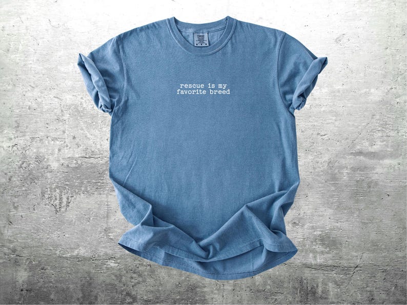 May include: A blue t-shirt with the phrase "rescue is my favorite breed" in white text. The shirt is made of a soft material and has rolled-up sleeves. The shirt is displayed on a gray, textured surface.