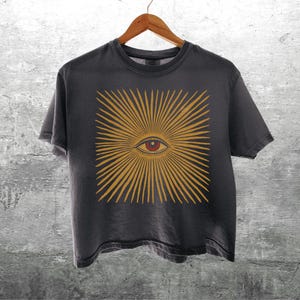 May include: A charcoal gray short-sleeve t-shirt featuring a central graphic of a stylized eye surrounded by radiating golden-yellow sunburst lines. The eye has a dark pupil, a red iris, and fine black line details. The shirt hangs on a wooden hanger.
