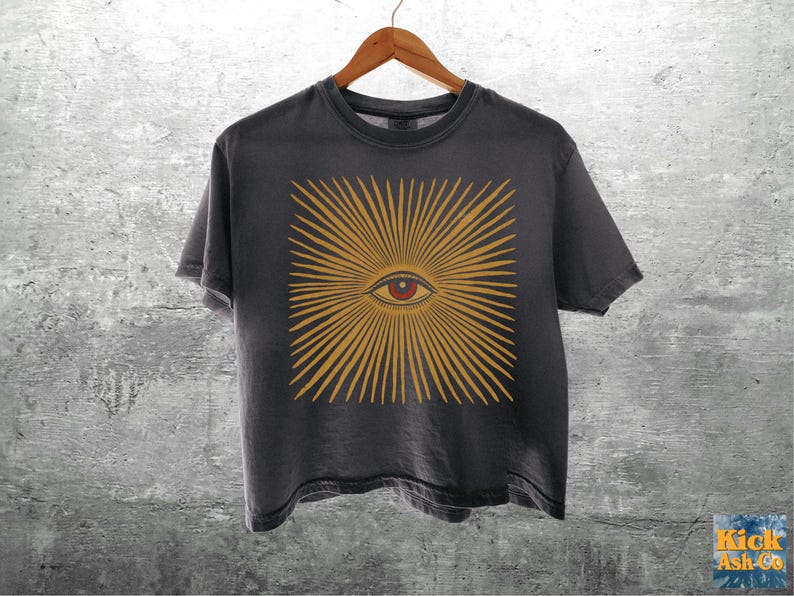 May include: A charcoal gray short-sleeve t-shirt featuring a central graphic of a stylized eye surrounded by radiating golden-yellow sunburst lines. The eye has a dark pupil, a red iris, and fine black line details. The shirt hangs on a wooden hanger.
