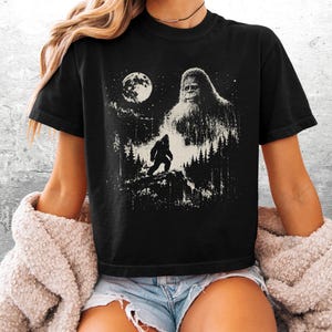 以下が含まれることがあります： 黒の半袖Tシャツ。満月の夜の森の中に立つ、ビッグフットに似た大きな毛むくじゃらの生き物が白のグラフィックでプリントされています。デザインには、尾根を歩く生き物の小さなシルエットも含まれています。