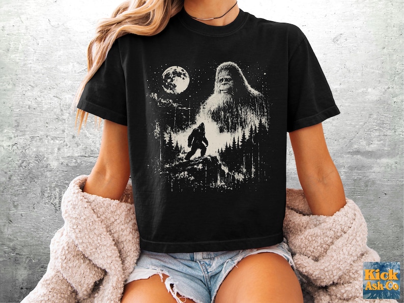 以下が含まれることがあります： 黒の半袖Tシャツ。満月の夜の森の中に立つ、ビッグフットに似た大きな毛むくじゃらの生き物が白のグラフィックでプリントされています。デザインには、尾根を歩く生き物の小さなシルエットも含まれています。