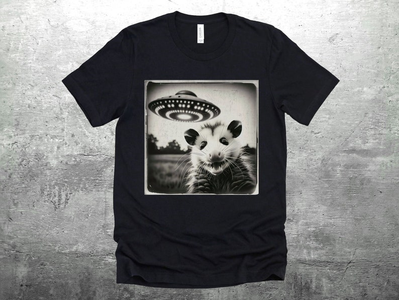 May include: Black and white graphic t-shirt featuring a photo of a possum looking up at a UFO. The photo is framed in a vintage style.