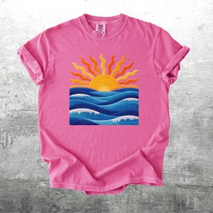 May include: A pink t-shirt featuring a colorful graphic of a sunset over ocean waves. The sun is yellow and orange, with blue waves below. The shirt has rolled-up sleeves.