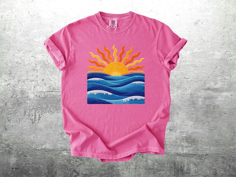 May include: A pink t-shirt featuring a colorful graphic of a sunset over ocean waves. The sun is yellow and orange, with blue waves below. The shirt has rolled-up sleeves.
