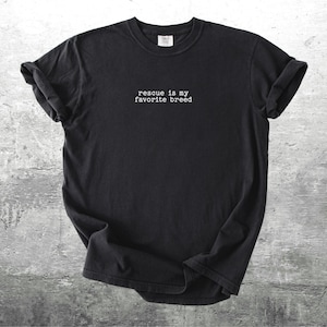 May include: Black t-shirt with the text "rescue is my favorite breed" in white, centered on the chest. The shirt has rolled-up sleeves and is displayed on a textured gray surface. A casual, comfortable style.