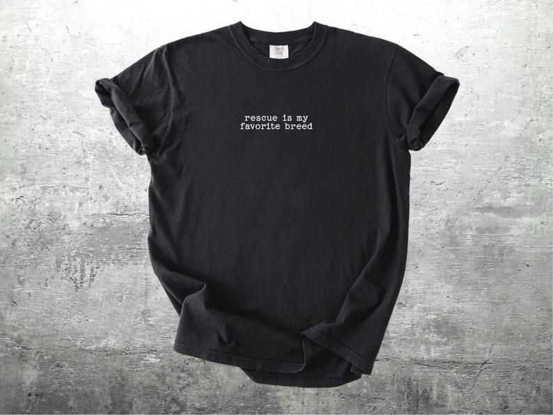 May include: Black t-shirt with the text "rescue is my favorite breed" in white, centered on the chest. The shirt has rolled-up sleeves and is displayed on a textured gray surface. A casual, comfortable style.