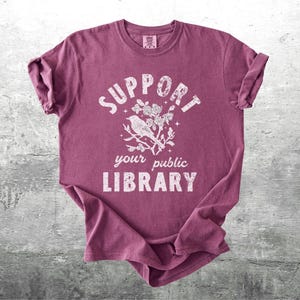 May include: A maroon t-shirt with rolled-up sleeves featuring the text "SUPPORT your public LIBRARY" in a distressed white font. A white illustration of a bird on a branch is centered above the words.