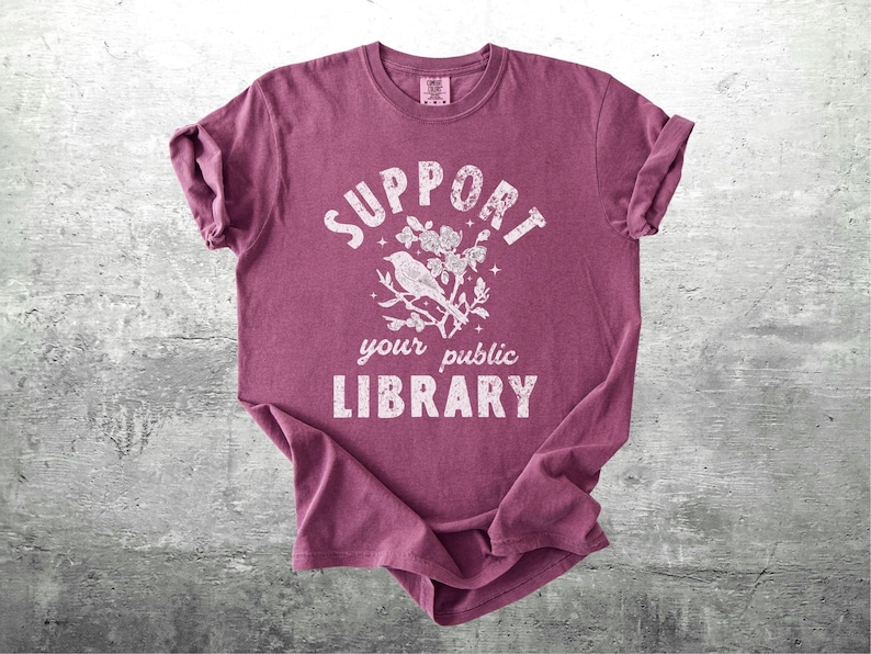 May include: A maroon t-shirt with rolled-up sleeves featuring the text "SUPPORT your public LIBRARY" in a distressed white font. A white illustration of a bird on a branch is centered above the words.