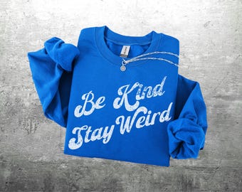 Puede incluir: Una sudadera azul real con las palabras "Be Kind Stay Weird" en letras blancas desgastadas. Un collar plateado con un pequeño colgante está sobre la camisa. La sudadera está doblada sobre una superficie gris.