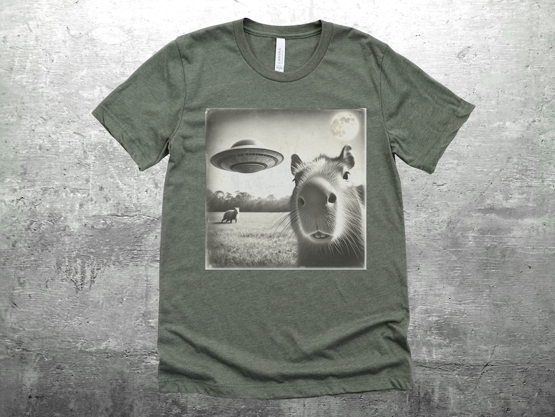K&ouml;nnte beinhalten: Ein gr&uuml;nes T-Shirt mit einem Schwarzwei&szlig;bild eines Capybaras, das zu einem UFO aufblickt. Das Bild zeigt auch ein kleines Tier im Hintergrund.