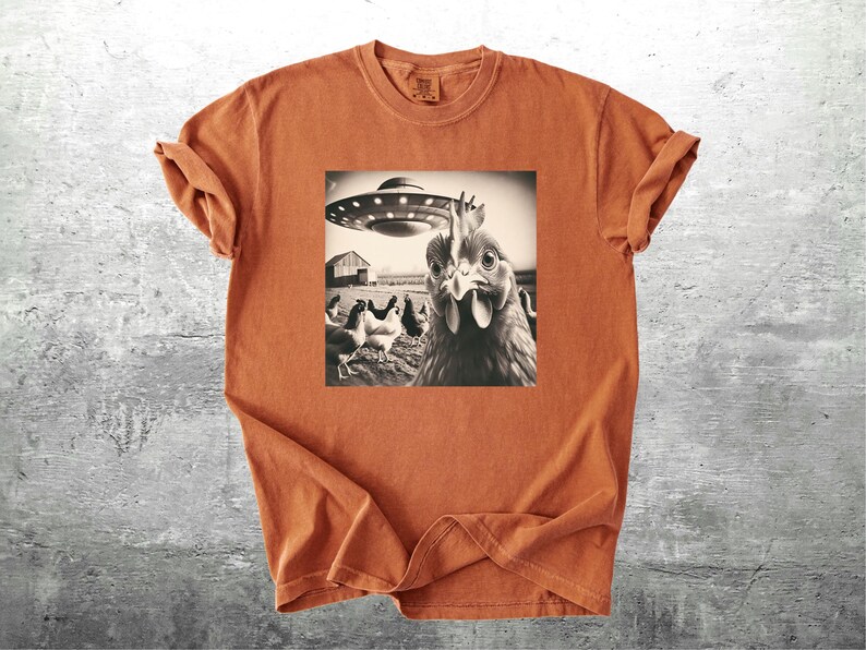 May include: Burnt orange t-shirt featuring a black and white graphic of a chicken looking at a UFO. Other chickens and a barn are in the background. The shirt has short sleeves and a crew neck.