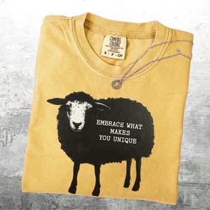 Puede incluir: Camiseta de color amarillo mostaza con un gr&aacute;fico de oveja negra y el texto "EMBRACE WHAT MAKES YOU UNIQUE". Un collar plateado con un peque&ntilde;o colgante descansa sobre la camiseta. La camiseta est&aacute; doblada.