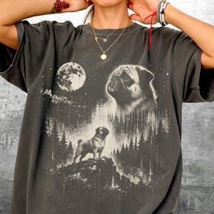 May include: Dark gray t-shirt featuring a pug design with a full moon and forest scene. The graphic includes a large pug face and a smaller pug standing on a hill. The text "Kick Ash Co" is in the lower right corner.