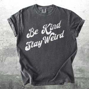 May include: Dark gray t-shirt with rolled-up sleeves. The shirt features the white text "Be Kind Stay Weird" in a casual, slightly distressed font. The shirt is made of a soft material and is likely pre-shrunk.