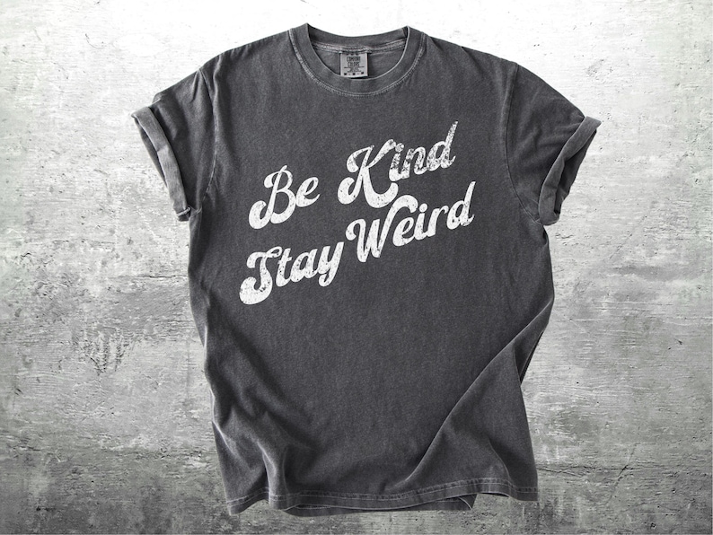 May include: Dark gray t-shirt with rolled-up sleeves. The shirt features the white text "Be Kind Stay Weird" in a casual, slightly distressed font. The shirt is made of a soft material and is likely pre-shrunk.
