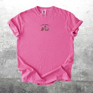 May include: A vibrant pink t-shirt with rolled sleeves. The shirt displays an embroidered design of a donkey and a box, with the text "I don't give a" above the image. The t-shirt is made of a soft material.