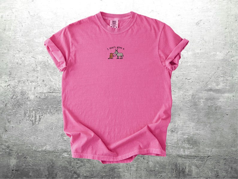 May include: A vibrant pink t-shirt with rolled sleeves. The shirt displays an embroidered design of a donkey and a box, with the text "I don't give a" above the image. The t-shirt is made of a soft material.