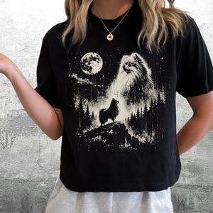 May include: A black short-sleeved t-shirt featuring a white graphic print of a fluffy dog standing on a rocky cliff in a forest at night. The design includes a large full moon, a starry sky, and a large ethereal dog face in the clouds above.