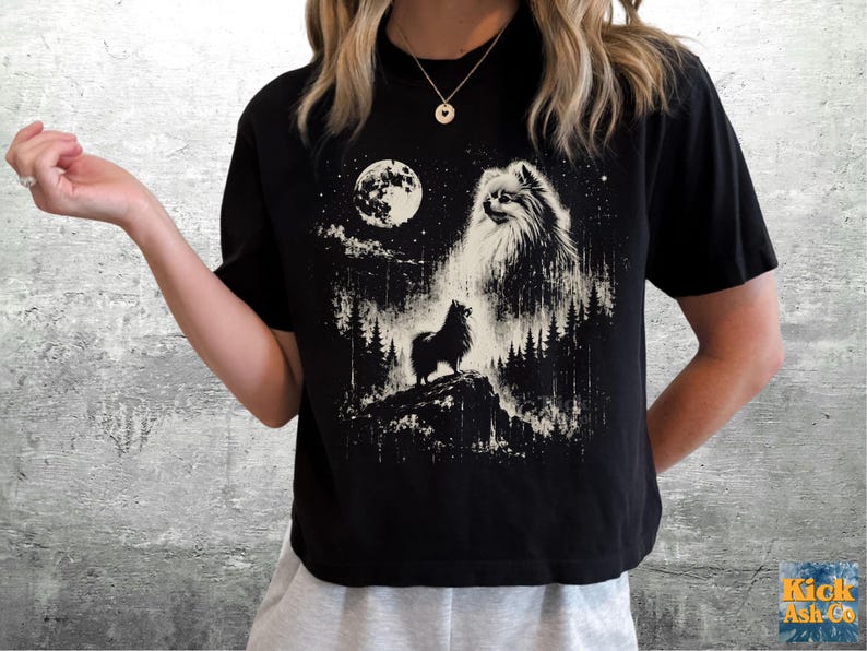 May include: A black short-sleeved t-shirt featuring a white graphic print of a fluffy dog standing on a rocky cliff in a forest at night. The design includes a large full moon, a starry sky, and a large ethereal dog face in the clouds above.