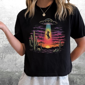 Puede incluir: Una camiseta negra de manga corta con un estampado gr&aacute;fico de un paisaje des&eacute;rtico al atardecer con cactus y un OVNI abduciendo a un perro peque&ntilde;o. El dise&ntilde;o utiliza colores vibrantes como el morado, rosa, naranja y azul para el cielo.