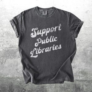 May include: Dark gray t-shirt with the words "Support Public Libraries" in white, stylized lettering. The shirt has a vintage, worn look, with rolled-up sleeves. The background is a textured, gray surface.