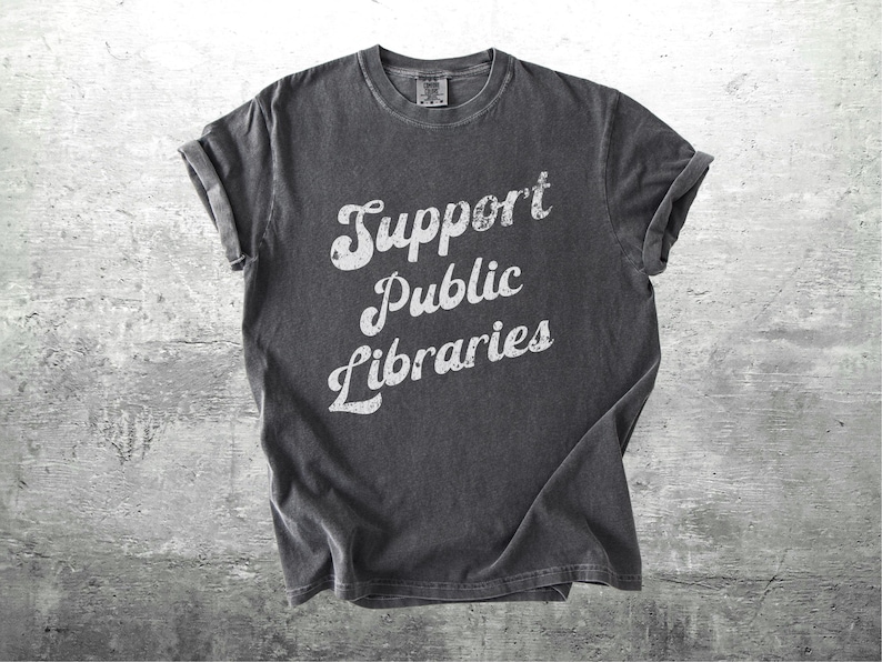 May include: Dark gray t-shirt with the words "Support Public Libraries" in white, stylized lettering. The shirt has a vintage, worn look, with rolled-up sleeves. The background is a textured, gray surface.