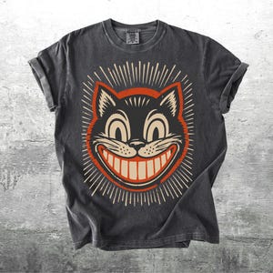 May include: A charcoal gray short-sleeve t-shirt featuring a graphic print of a black cat face with a wide, toothy grin, large eyes, and radiating lines in cream and orange colors. The design has a distressed, vintage-inspired aesthetic.