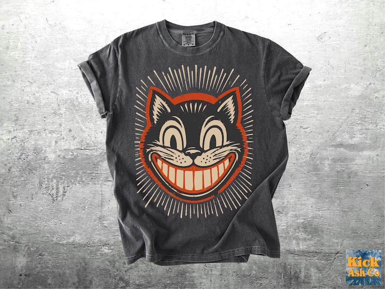 Op de afbeelding: Een antracietgrijs T-shirt met korte mouwen en een grafische print van een zwart kattengezicht met een brede, tandige grijns, grote ogen en stralende lijnen in cr&egrave;me en oranje. Het ontwerp heeft een verweerde, vintage uitstraling.
