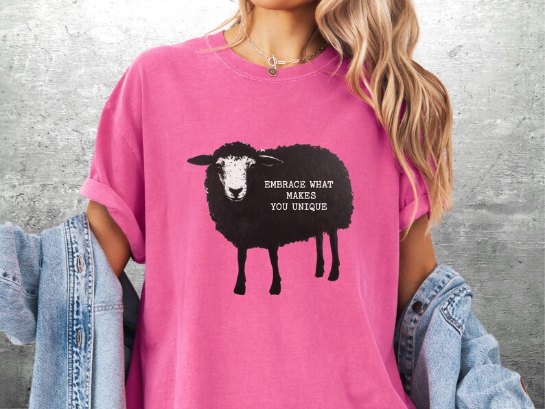 Puede incluir: Camiseta rosa brillante con un dise&ntilde;o de oveja negra y el texto "EMBRACE WHAT MAKES YOU UNIQUE". La camiseta tiene mangas cortas y un corte holgado, ideal para uso casual.