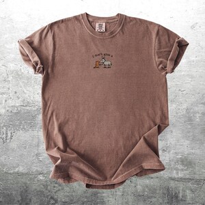 May include: A brown t-shirt with rolled sleeves. The shirt has the embroidered phrase "I don't give a" above a cartoon donkey and a box. The t-shirt is made of a soft material and has a vintage appearance.