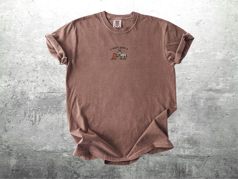 May include: A brown t-shirt with rolled sleeves. The shirt has the embroidered phrase "I don't give a" above a cartoon donkey and a box. The t-shirt is made of a soft material and has a vintage appearance.