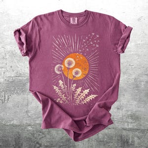 May include: A maroon t-shirt featuring a graphic of dandelions in front of an orange sunburst. The design includes white radiating lines and dandelion seeds. The shirt is made of soft material and has rolled-up sleeves.