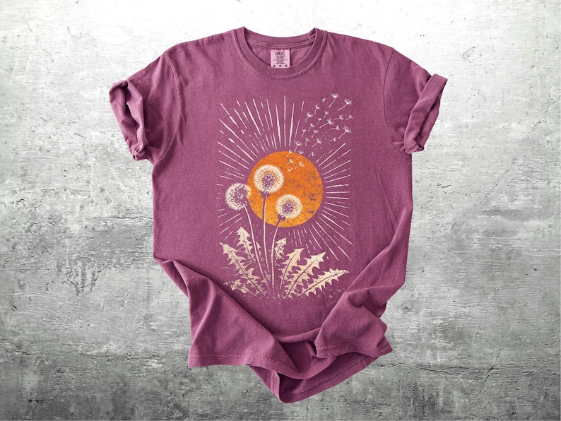 May include: A maroon t-shirt featuring a graphic of dandelions in front of an orange sunburst. The design includes white radiating lines and dandelion seeds. The shirt is made of soft material and has rolled-up sleeves.