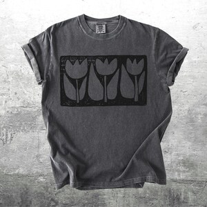 May include: Dark gray t-shirt with a black graphic of three tulips in a rectangular frame. The shirt has rolled-up sleeves and a small tag at the neckline. The background is a textured, light gray surface.