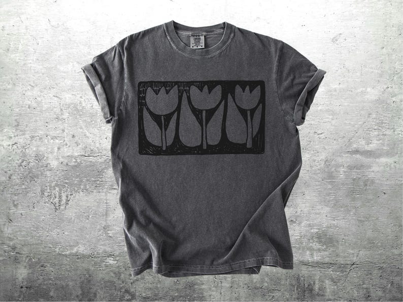 May include: Dark gray t-shirt with a black graphic of three tulips in a rectangular frame. The shirt has rolled-up sleeves and a small tag at the neckline. The background is a textured, light gray surface.