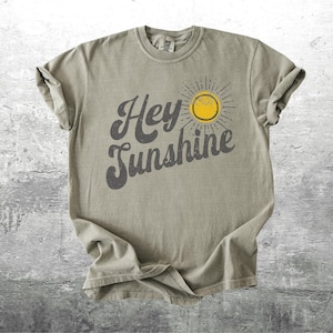 May include: A light olive green t-shirt with the words "Hey Sunshine" in a dark gray, stylized font. A yellow sun graphic with gray rays is to the right of the text. The shirt is made of a soft material.