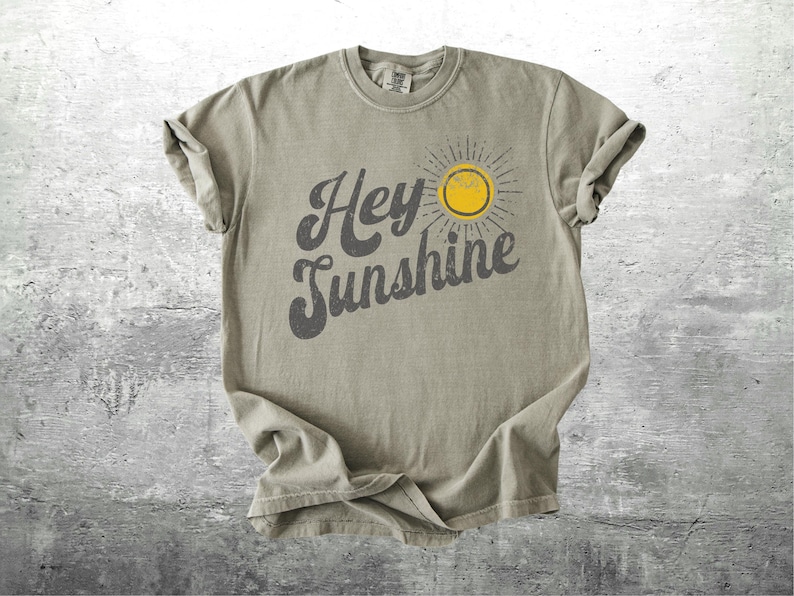 May include: A light olive green t-shirt with the words "Hey Sunshine" in a dark gray, stylized font. A yellow sun graphic with gray rays is to the right of the text. The shirt is made of a soft material.