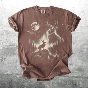 May include: Brown t-shirt featuring a white graphic of a jackalope with antlers, a full moon, and a forest scene. The design includes a jackalope on a hilltop and another in the forest. The shirt has a crew neck and short sleeves.