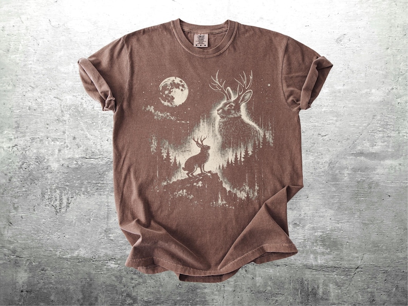 May include: Brown t-shirt featuring a white graphic of a jackalope with antlers, a full moon, and a forest scene. The design includes a jackalope on a hilltop and another in the forest. The shirt has a crew neck and short sleeves.
